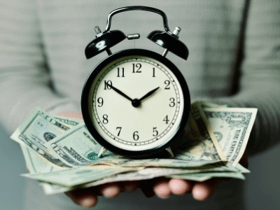 Imagen de un reloj sobre una pila de dinero, ambos sostenidos por unas manos. Simboliza el alto valor de una casa en el norte de Mérida